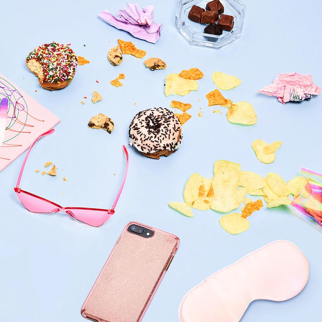 Donuts, chips and various junk food spread on a white surface alongside sunglasses, cell phone and eye mask