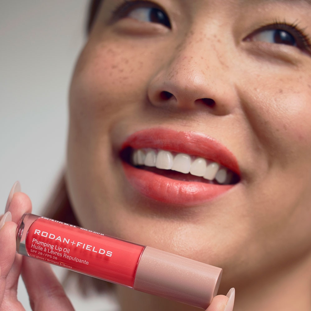 Young woman holding Rodan + Fields Plumping Lip Oil SPF 28 in the Coral shade, showing her lips after applying the product.